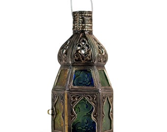 May include: An ornate, silver-colored lantern with a clear handle. The lantern features intricate filigree designs and stained glass panels in shades of green and blue. The base and top have decorative patterns.