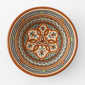 May include: A circular ceramic plate with an intricate pattern. The design includes a central floral element in orange and cream, encircled by bands of black, green, and brown. The rim is a warm brown colour, and the plate has a detailed, symmetrical design.