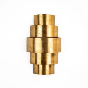 Könnte beinhalten: Eine goldfarbene, gestapelte zylindrische Skulptur vor weißem Hintergrund. Die Skulptur besteht aus vier Zylindern unterschiedlicher Größe, wodurch ein gestuftes Aussehen entsteht. Das Metall hat ein leicht gealtertes Aussehen.