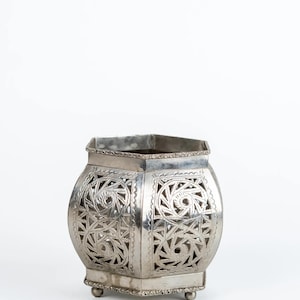 May include: A silver-colored, decorative lantern with an openwork design. The lantern has a geometric shape with intricate patterns and sits on small, rounded feet. The top edge has a decorative border.