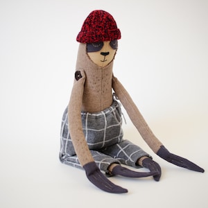 Könnte beinhalten: Eine Faultier-Stoffpuppe mit beigem Stoffkörper, die eine rot-schwarze Strickmütze, dunkelgraue Augenpartien und eine graue Hose mit weißem Karomuster trägt. Die Puppe hat lange Arme und Beine mit Händen und Füßen aus dunkelgrauem Stoff.