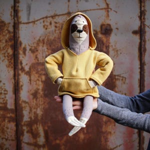 Op de afbeelding: Een luiaardknuffel met een lichtgrijs lichaam, donkerbruine oogvlekken en een gestikte neus. De luiaard draagt een felgele hoodie met een kangoeroezak aan de voorzijde en lange mouwen, met de armen in de zij geplaatst.