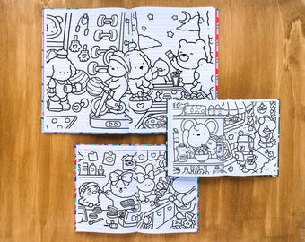 May include: Three open coloring books with black and white illustrations of cartoon characters in various settings. One book shows a gym scene, another a cafe, and the third a kitchen. The books are on a wooden surface.