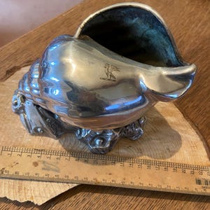 May include: A silver-coloured, seashell-shaped decorative container. The container has a detailed, textured surface and a dark interior. A small emblem is visible on the side. The container is resting on a wooden surface.
