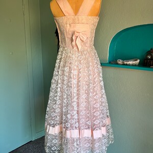 May include: A pink lace dress with a satin bow on the back. The dress has a fitted bodice and a full skirt. The skirt has a band of satin ribbon around the hem.