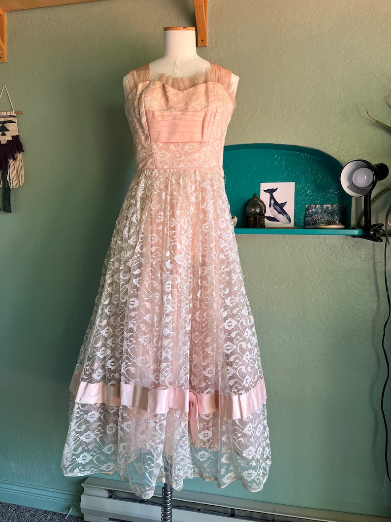 May include: A vintage pink and white lace dress with a sweetheart neckline and a full skirt. The dress has a satin ribbon at the waist and a layered skirt.