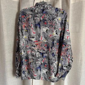 May include: A button-down shirt with a black and white abstract pattern, accented with red, blue, and yellow details. The long-sleeved shirt is hanging on a hanger against a neutral background.