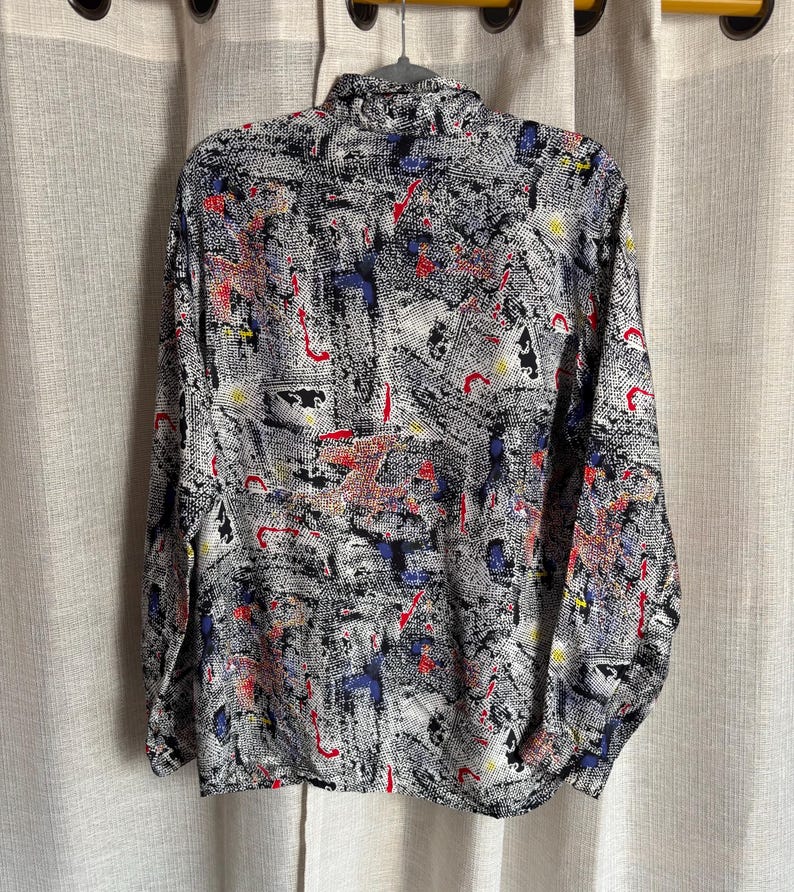 May include: A button-down shirt with a black and white abstract pattern, accented with red, blue, and yellow details. The long-sleeved shirt is hanging on a hanger against a neutral background.