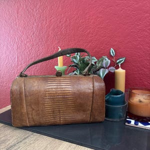 Könnte beinhalten: Eine rechteckige braune Handtasche mit texturiertem Eidechsenhaut-Muster, ausgestattet mit einem einzelnen gebogenen Tragegriff, einem Metallrahmen und einem zentralen runden Verschluss. Die Tasche hat eine strukturierte Form mit glatten Seitenteilen.