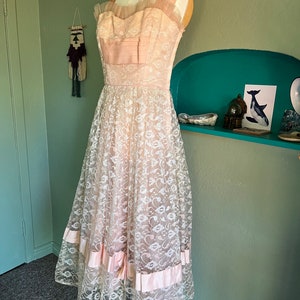 May include: A vintage, sleeveless, pink and white lace dress with a sweetheart neckline and a full skirt. The dress has a pink satin ribbon at the waist and at the hem.