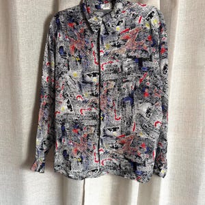 May include: A patterned button-down shirt in black, white, red, blue, and yellow. The long-sleeved shirt is hanging on a grey hanger. The shirt has a collared neckline and a pocket on the left chest.