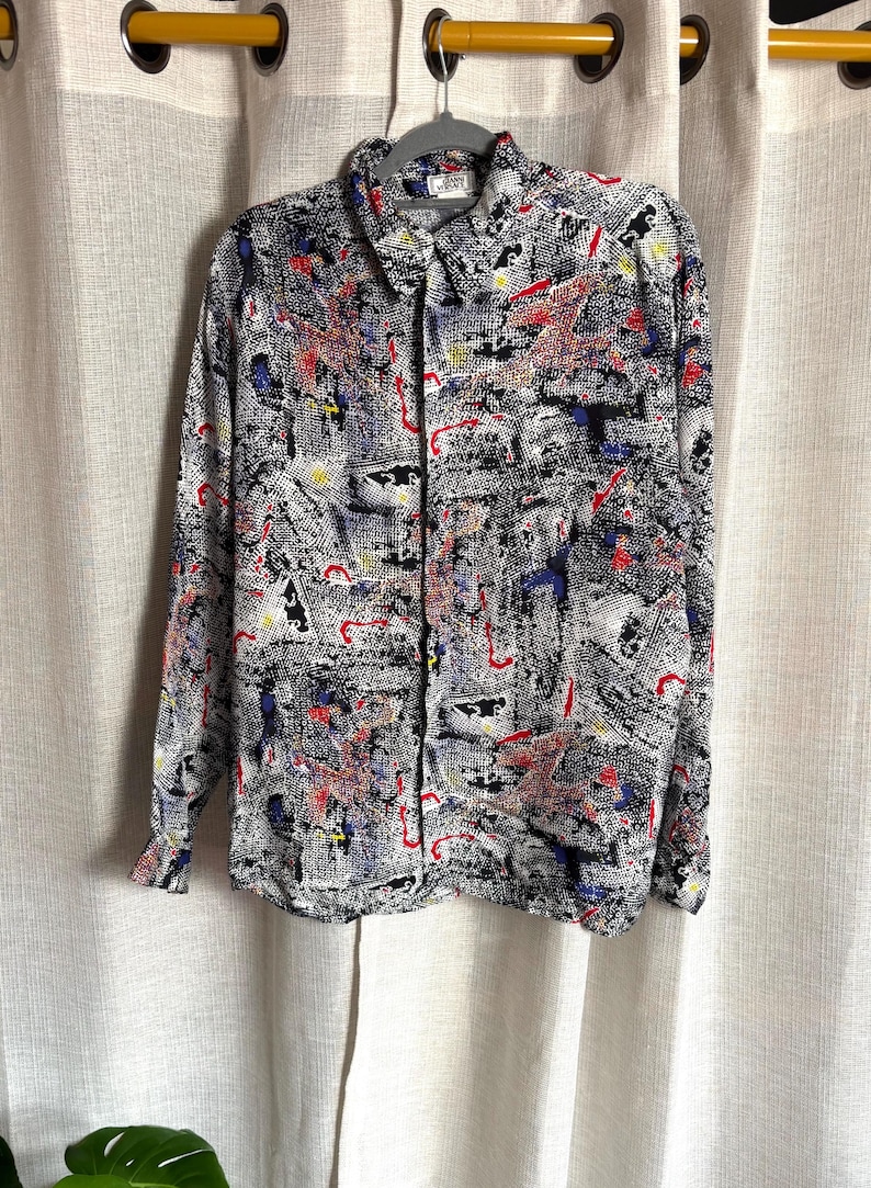 May include: Button-down shirt with a black, white, red, blue, and yellow abstract pattern. The long-sleeved shirt is hanging on a gray hanger. The shirt has a collared neckline and a pocket on the left chest.