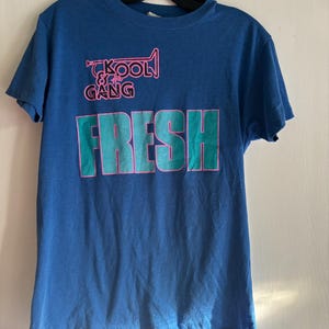May include: A blue short-sleeved t-shirt featuring a graphic print in pink and teal. The design includes a pink trumpet icon above the text "KOOL & the GANG" and the word "FRESH" printed in large teal block letters with a pink outline.