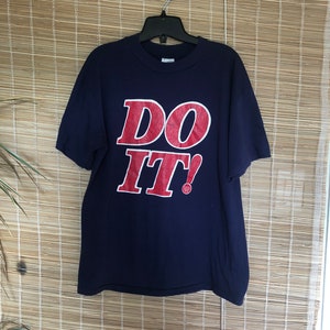 May include: A navy blue t-shirt displaying the words "DO IT!" in red with a white outline. The tee is suspended on a black hanger. The background is a bamboo blind. A simple, striking design.