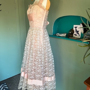 May include: A vintage pink lace dress with a sweetheart neckline and a full skirt. The dress has a pink satin ribbon at the waist and a pink satin trim at the hem.