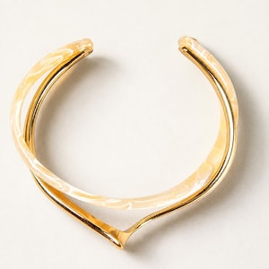 May include: A cuff bracelet featuring a polished gold-tone metal band with a V-shaped centre point, layered with a smooth, translucent cream and white marbled resin band that follows a similar curved silhouette in an open design.