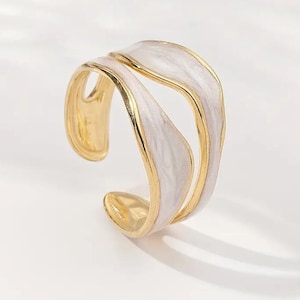 May include: An open-style gold ring featuring two wavy, parallel bands. Each band is inlaid with a smooth, pearlescent white enamel finish that follows the organic, flowing curves of the metal structure, creating a layered and dimensional appearance.