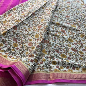 May include: A textile featuring a cream-colored base with a dense floral pattern in shades of yellow, orange, and dark green. The fabric includes a wide, vibrant magenta border with intricate gold-toned metallic woven detailing.