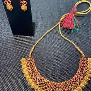 May include: A gold-coloured necklace and matching earrings set. The necklace has a curved design with many red gemstones. The earrings are small, with a similar design. A yellow cord with red and green tassels is also visible.