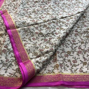 May include: A cream-colored fabric featuring a repeating floral vine pattern in shades of brown, red, and green. The textile is bordered by a vibrant magenta band with intricate gold-toned metallic thread detailing along the edges.