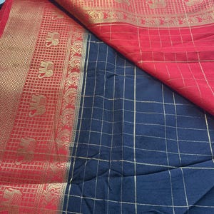 May include: A folded textile featuring a vibrant red and gold border with elephant motifs. The fabric transitions to a navy blue checkered pattern with gold lines, creating a striking contrast. The textile appears to be a saree.