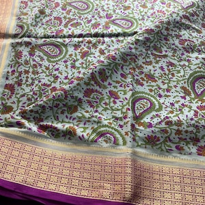 May include: A white fabric textile featuring a repeating floral and paisley pattern in shades of green, purple, and orange. The fabric is bordered by a wide, deep purple band with a metallic gold geometric weave and a thin gold trim.