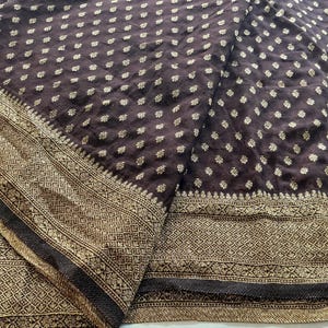May include: A dark brown fabric featuring a repeating pattern of small gold floral motifs. The textile includes a wide, ornate border with intricate gold geometric and floral designs, along with a section displaying thin, vertical white stripes.
