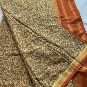 May include: A beige fabric featuring a dense, repeating pattern of small brown paisley and floral motifs. The textile includes a wide, solid burnt orange border along one side, accented by thin, metallic gold horizontal stripes.