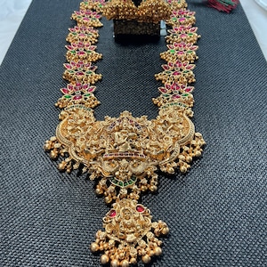 May include: A gold-colored necklace and matching earrings set. The necklace features intricate detailing with red and green accents. The design includes a central pendant with a detailed figure and dangling gold beads. The earrings have a similar design.