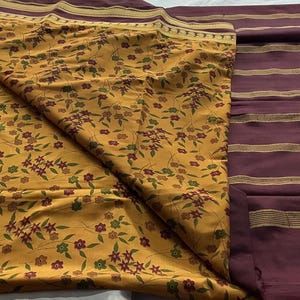May include: A textile fabric featuring a mustard yellow section with a small floral pattern in red and green, contrasted by a deep plum purple section with horizontal gold stripes and a decorative gold border along the edges of the material.
