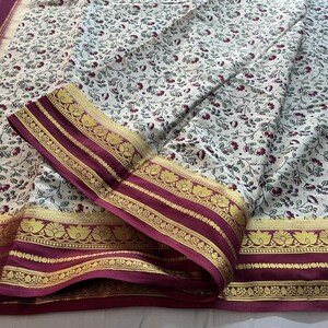 May include: A cream-colored fabric featuring a repeating pattern of small dark red flowers and green leaves. The textile is bordered by a wide, deep maroon band adorned with intricate gold-toned floral and geometric woven designs.