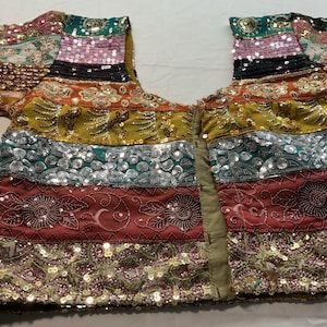 Puede incluir: Una colorida blusa adornada con mangas cortas. La prenda presenta bandas horizontales de varios colores, incluyendo rosa, dorado, turquesa y rojo, adornadas con lentejuelas y bordados intrincados. La blusa tiene un escote en V y un cierre central.