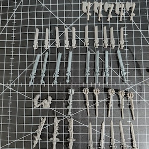 Könnte beinhalten: Eine Sammlung unbemalter, grauer Miniaturwaffen aus Kunststoff, darunter Kettenschwerter, Äxte, Schwerter und Streitkolben, angeordnet auf einer schwarzen Schneidematte. Wahrscheinlich für Tabletop-Spiele oder Modellbau.
