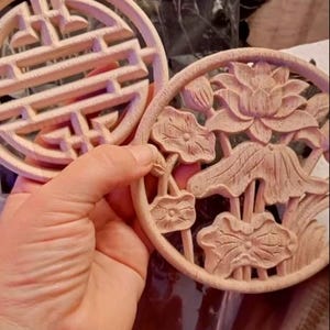 May include: Two round, wooden coasters with intricate designs. One coaster features a geometric pattern, while the other has a floral design with carved lotus flowers.