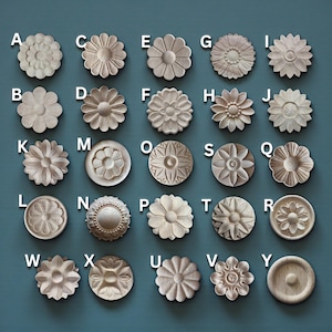 May include: A selection of twenty-five wooden floral embellishments, each with a unique carved design. The decorative elements are arranged in a grid and labelled with letters A to Y. The carvings vary in style, with some resembling daisies and others more abstract floral patterns.
