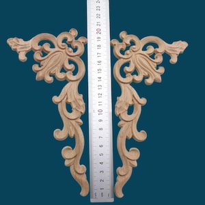 以下が含まれることがあります： 複雑な花の模様が施された、未完成の木製のコーナーブラケット2個。ブラケットは高さ約17cmで、定規の横に置かれています。