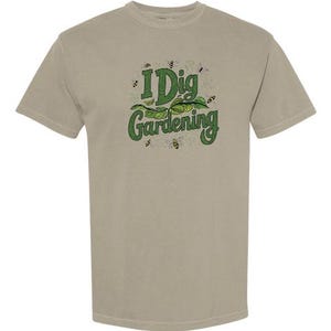 May include: A tan-colored t-shirt with the text "I Dig Gardening" in green, with a leaf and bee graphic. The shirt is made of a soft material and has a crew neck.
