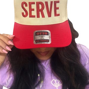 Könnte beinhalten: Beige und rote Baseballkappe mit dem Wort "SERVE" in Rot gestickt. Die Kappe hat eine klassische Passform und ein mittleres Profil. Der Hut ist aus hochwertigem Stoff gefertigt und ein beliebter Stil.