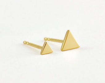 May include: Two gold-colored triangle stud earrings. One earring is smaller than the other. The earrings are on a white background. The earrings are likely made of gold.
