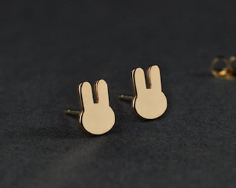 May include: Gold-coloured bunny stud earrings. The earrings have a simple, minimalist bunny silhouette design. The image also shows earring backs. The background is a dark grey colour.