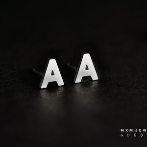 May include: A pair of silver-colored letter "A" stud earrings. The earrings are displayed on a dark, textured surface. The earrings are simple and modern, with a minimalist design. The brand name "MXM JEWELRY & DESIGN" is at the bottom.