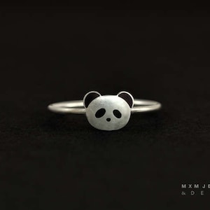 May include: A silver ring with a panda bear face design. The panda's eyes and ears are black.