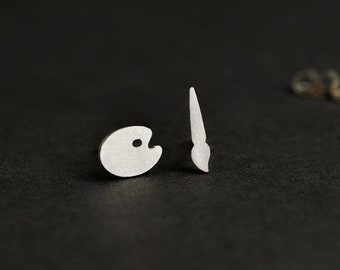 May include: A pair of silver earrings, one shaped like a paintbrush and the other like a paint palette.