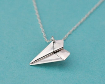 May include: A silver necklace with a paper airplane pendant. The necklace is on a light blue background.