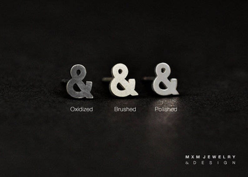 Puede incluir: Tres pendientes de plata con forma de ampersand en diferentes acabados: oxidado, cepillado y pulido.
