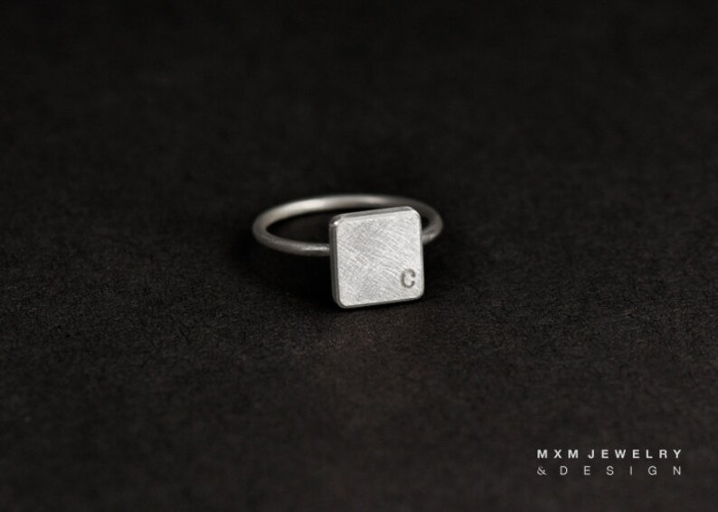 Puede incluir: Un anillo de plata con una banda de forma cuadrada con un acabado cepillado y la letra "C" grabada en la parte superior.