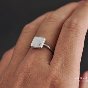 Puede incluir: Un anillo de plata con una banda de forma cuadrada que presenta la letra "C" grabada en la parte superior.