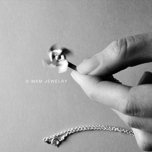 May include: A silver necklace with a spinning propeller charm. The charm is in motion and appears to be spinning. The necklace is on a chain and is being held by a hand.