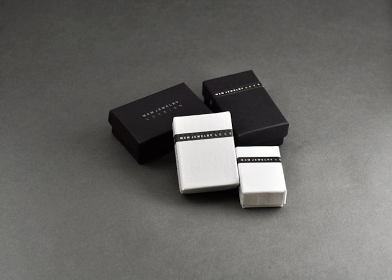 Puede incluir: Cuatro peque&ntilde;as cajas rectangulares, dos negras y dos blancas, con el texto "MWM JEWELRY EDITION" impreso en las cajas negras y "MWM JEWELRY & DESIGN" impreso en las cajas blancas.