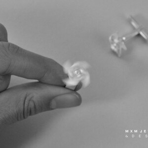 Puede incluir: Un par de gemelos de plata con un dise&ntilde;o giratorio. Los gemelos se muestran sobre una superficie blanca. El texto "MXM JEWELRY & DESIGN" es visible en la esquina inferior derecha de la imagen.